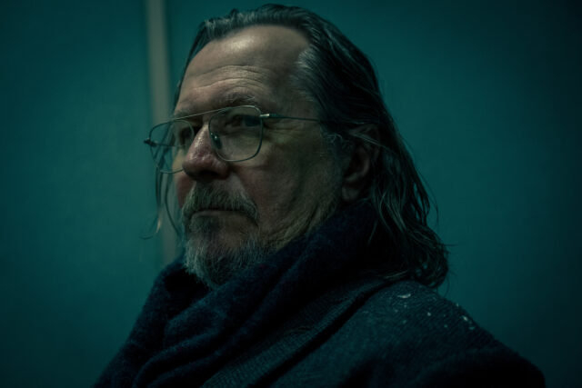 A close up of Gary Oldman with long, slicked-back hair and a grey beard, wearing large glasses and a heavy dark scarf, set against a moody teal backdrop. His expression is serious and contemplative, and the dim, shadowy lighting adds a gritty, noir-like atmosphere to the portrait.